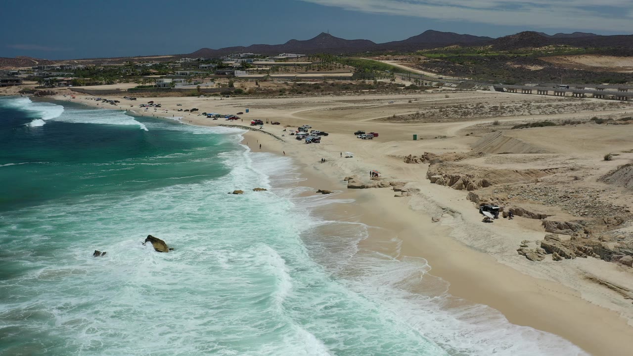 Wide drone view of waves, sand, rocky shore, beach vehicles, and desert hills.