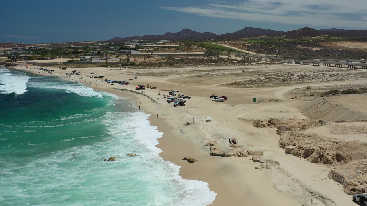 A high view over Los Cabos, Mexico shows turquoise waves at left, a sandy beach with scattered parked vehicles, dirt roads, low buildings, and distant mountains under bright light.