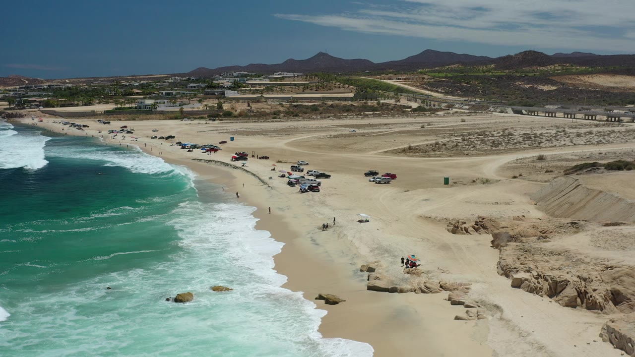 Aerial coastline view with green-blue water, sandy shore, beach vehicles, and rocky foreground edge.