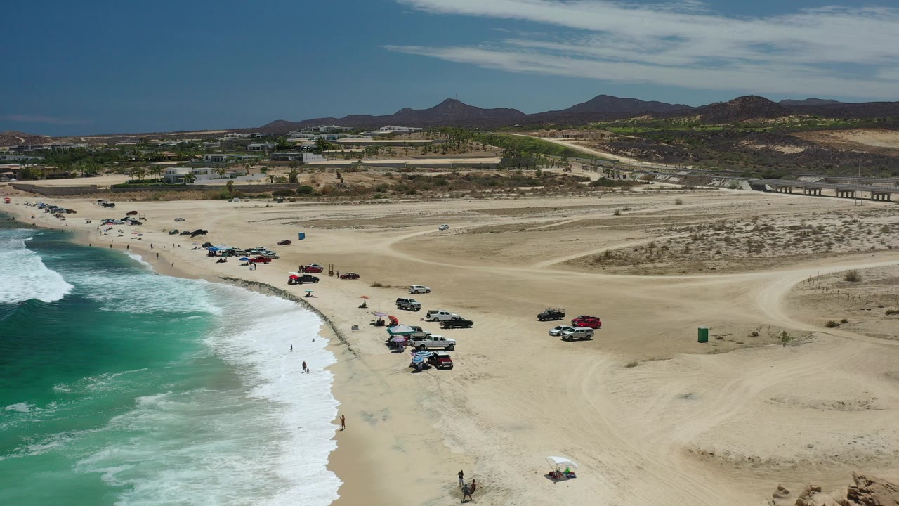 Aerial view of surf, sandy beach, parked vehicles, and distant hills.
