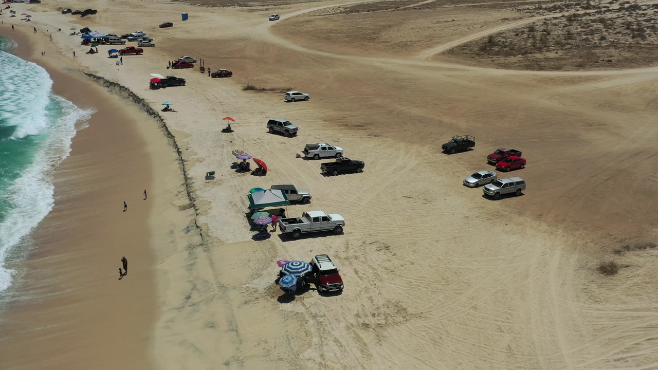 A high aerial frame over a Los Cabos beach shows surf at left, a line of wet sand, parked trucks and cars, shade umbrellas, a square tent, and small people near the shoreline.