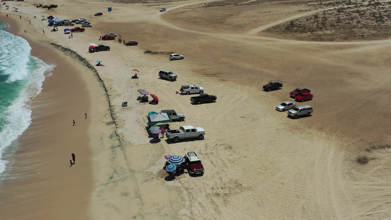 The next aerial frame holds over the Los Cabos shoreline with white surf rolling in, vehicles spaced along the beach, umbrellas near the water, and open tan sand extending inland.