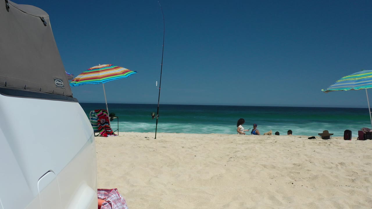 Beach scene with vehicle foreground, striped umbrellas, fishing rods, surf, and small people near the water.