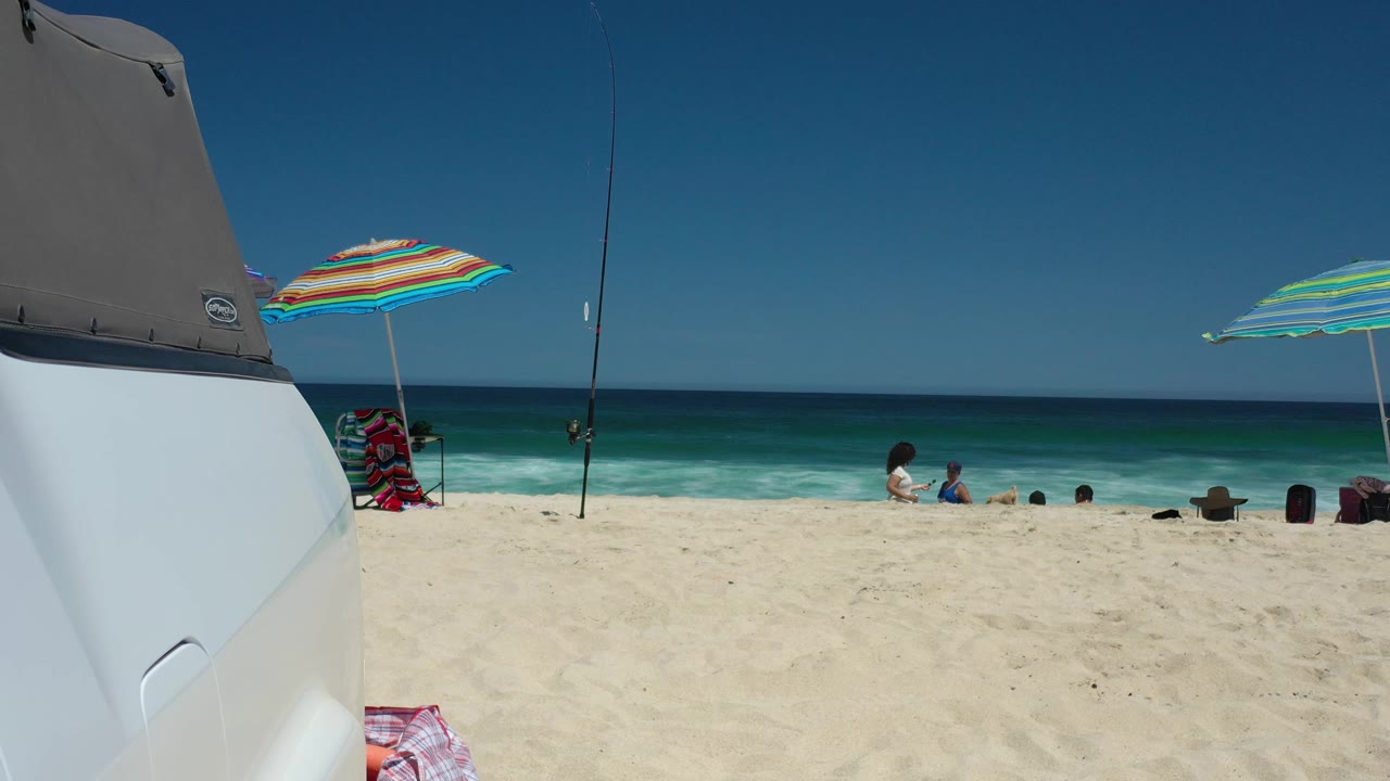Los Cabos shoreline frame with the vehicle still at left, a multicolor umbrella near center, fishing rods against the sky, and beachgoers sitting farther right by the water.
