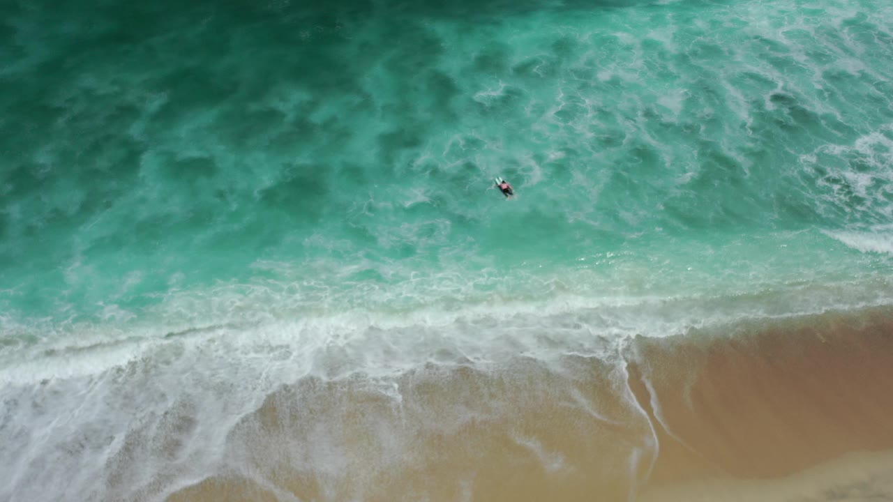 Overhead Los Cabos beach frame with turquoise water, white foam, tan sand, and a small dark object offshore, with no clearly visible people.