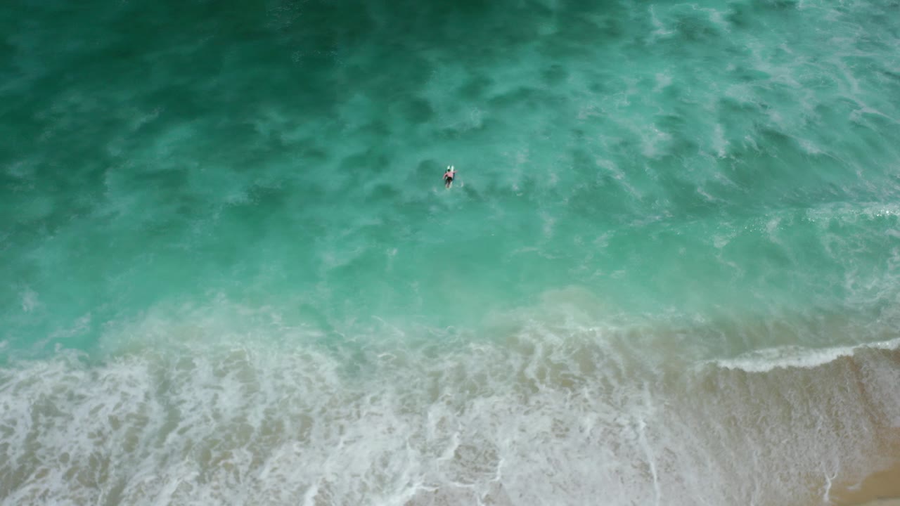 Overhead view of a surfer in turquoise surf near a sandy beach.