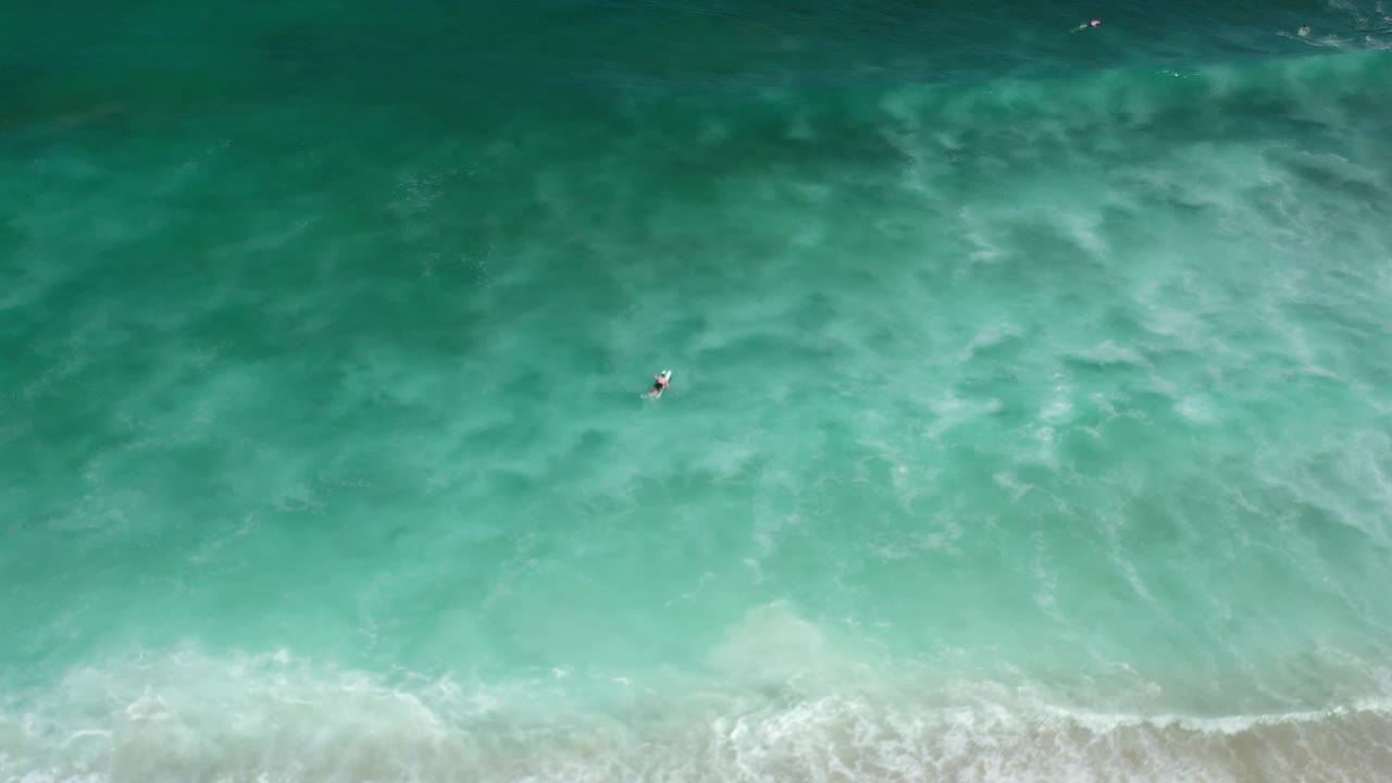 Overhead Los Cabos ocean frame with the surfer slightly left of center, surrounded by teal water gradients and a soft band of shoreward foam at the bottom.