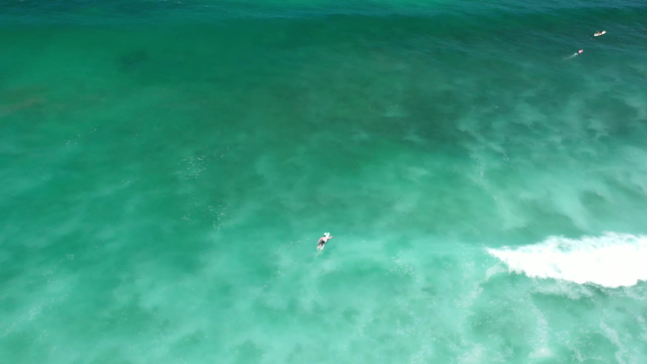Aerial Los Cabos water frame with a small surfer left of center, white surf spreading to the right, and darker green water near the top.