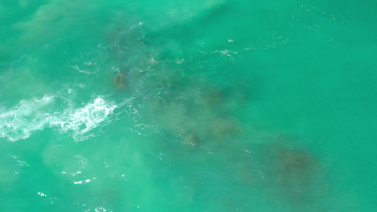Overhead view of turquoise water in Los Cabos, Mexico, with white foam breaking near darker submerged rocks and no visible people.
