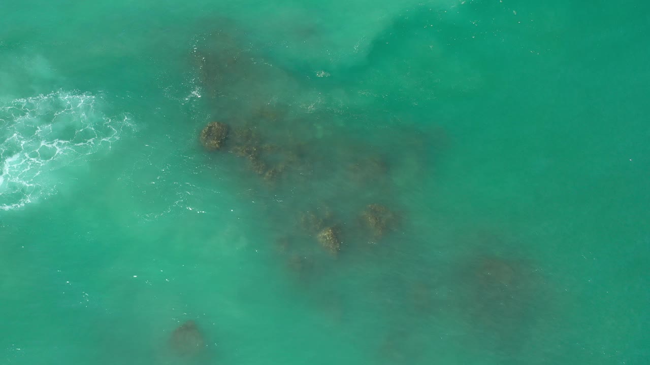 Aquamarine water in Los Cabos covers dark submerged reef or rocks, with a white foam patch entering from the left and no visible people.