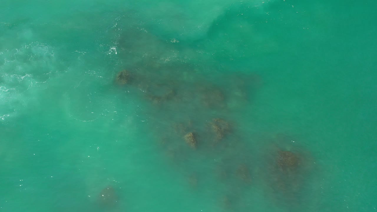Overhead view of turquoise water and dark submerged rocks in Los Cabos, Mexico, with small white wave breaks near the upper left and no visible people.