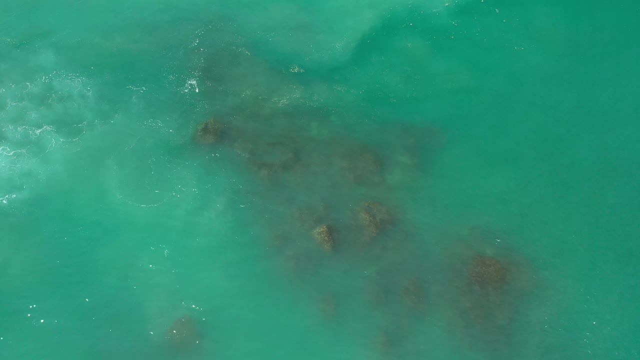 Aerial frame of green sea over scattered dark rocks in Los Cabos, Mexico, with faint white foam lines and no visible people.
