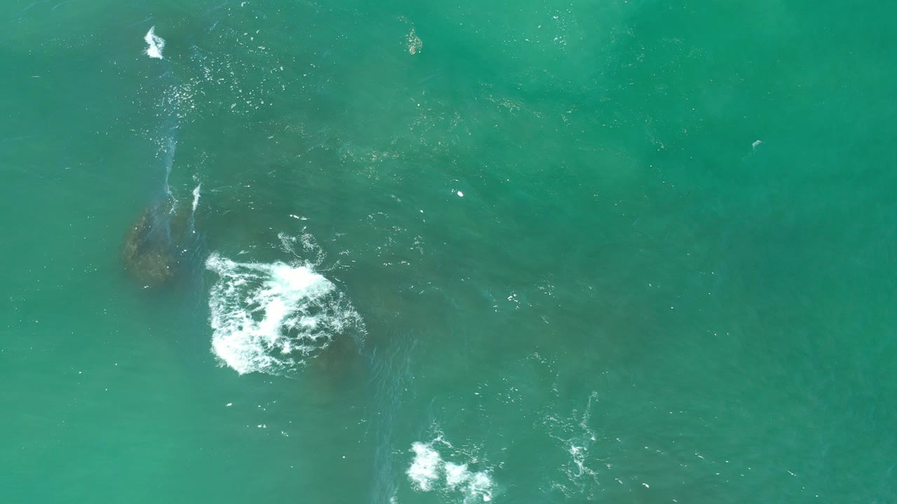 Overhead view of green ocean water in Los Cabos, Mexico, with white foam breaking near submerged dark rocks and no visible people.