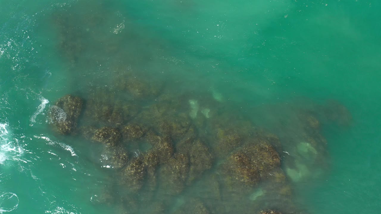 Overhead view of turquoise water and dark submerged rocks in Los Cabos, Mexico, with small foam traces and no visible people.