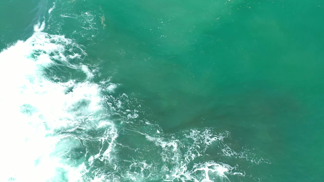 Overhead Los Cabos water frame where white foam spreads farther across green-blue ocean, with darker underwater tones and no visible people.