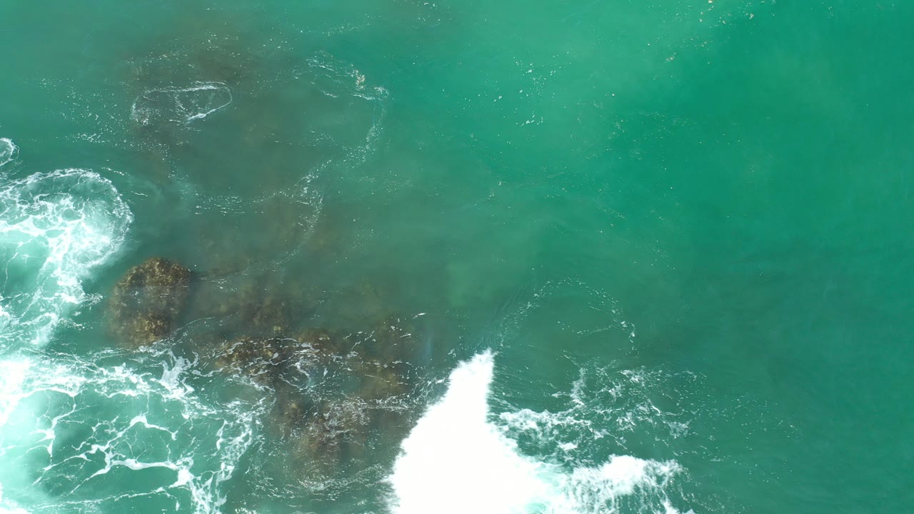 Overhead view of turquoise water in Los Cabos, Mexico, with white foam breaking around dark submerged patches near the lower edge and no visible people.
