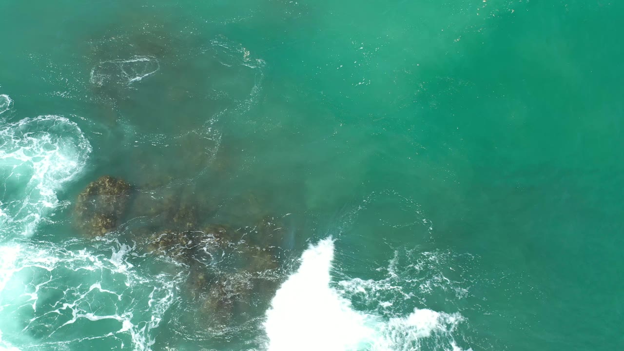 Aerial Los Cabos water view with surf foam widening over dark nearshore patches, green-blue water filling the frame, and no visible people.