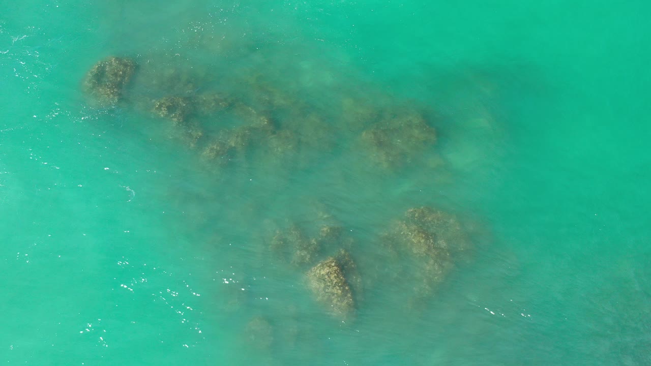 Overhead view in Los Cabos, Mexico of turquoise seawater with several dark submerged rock patches near the upper half and no visible people.