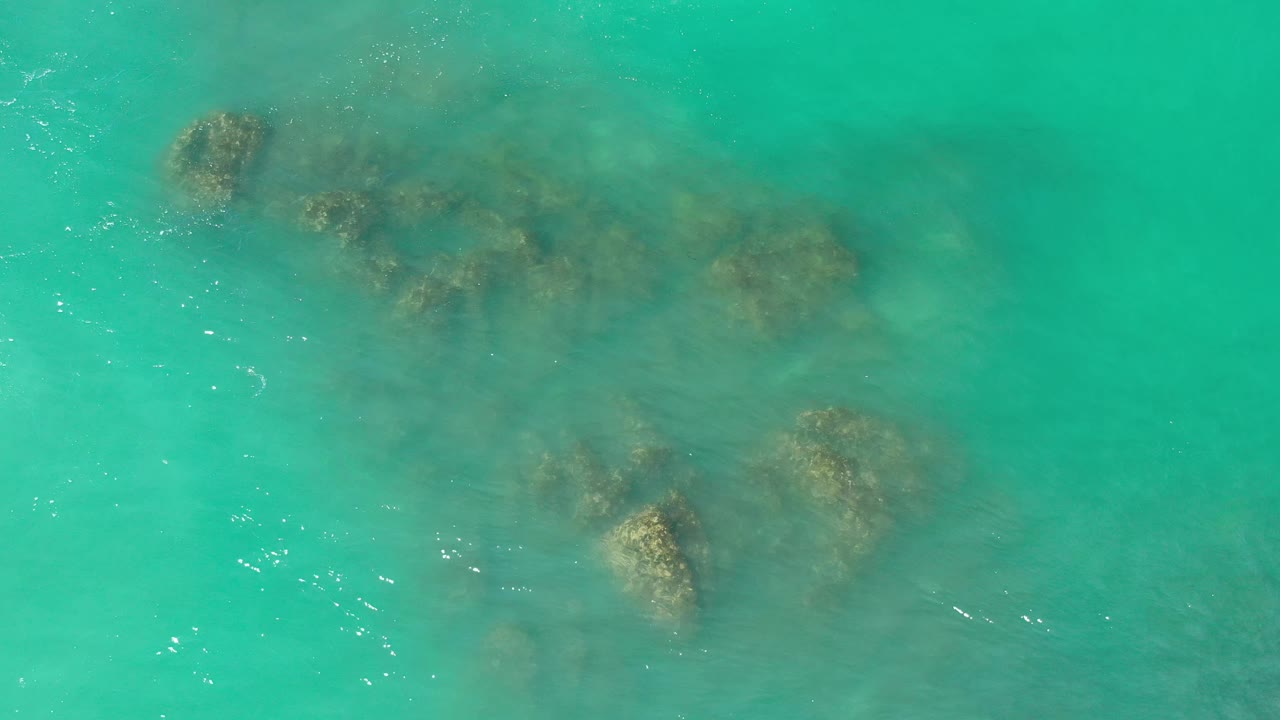 Overhead Los Cabos water view with darker rock shapes running diagonally through clear turquoise sea and no visible people.