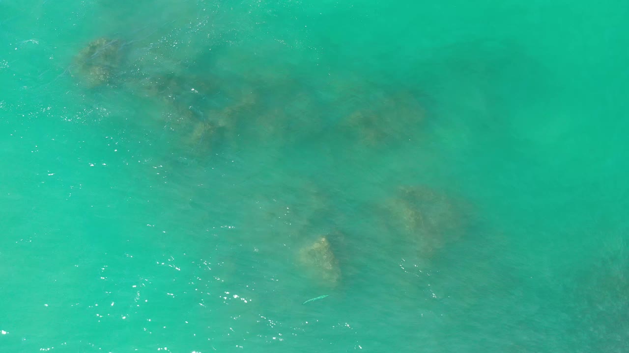 Overhead Los Cabos water view with faint brown reef shapes spread beneath green-blue shallows, scattered surface sparkle, and no visible people.