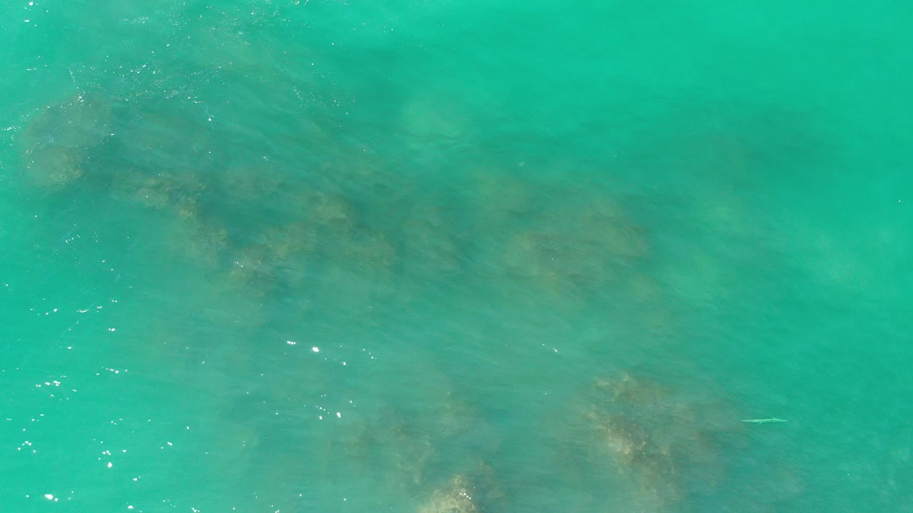 Top-down view of turquoise water and faint submerged rocks in Los Cabos, Mexico, with small surface glints and no visible people.