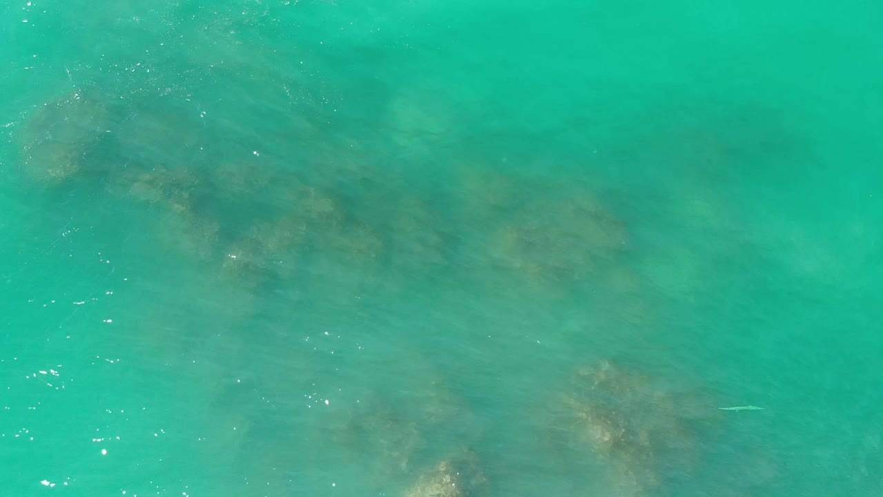 Overhead Los Cabos, Mexico frame showing blue-green water, muted rock forms beneath the surface, and no visible people.