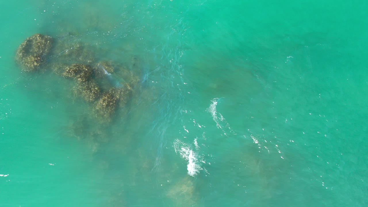 Overhead view of turquoise water in Los Cabos, Mexico, with several dark reef rocks beneath the surface, scattered white foam, and no visible people.