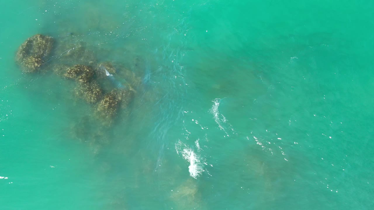 Aerial frame from Los Cabos, Mexico, showing a compact reef cluster under green-blue water, faint surface ripples, and no visible people.