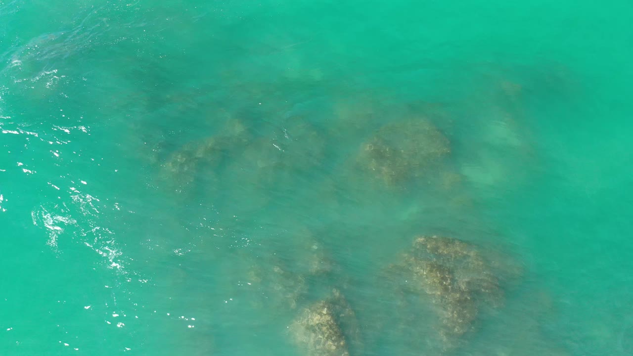 Aerial-looking frame of blue-green Los Cabos water with darker submerged rocks spread across the center and no visible people.