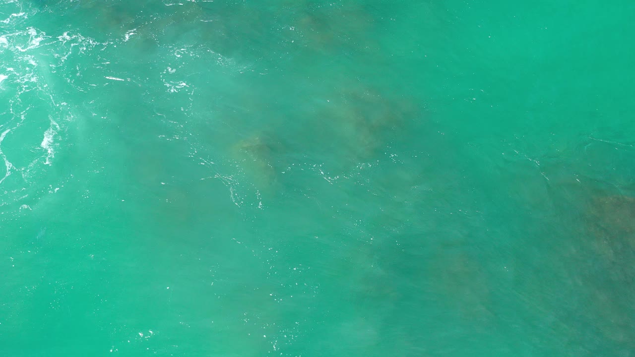 Overhead view of turquoise water in Los Cabos with faint submerged rock shapes, small white sun glints, and no visible people.