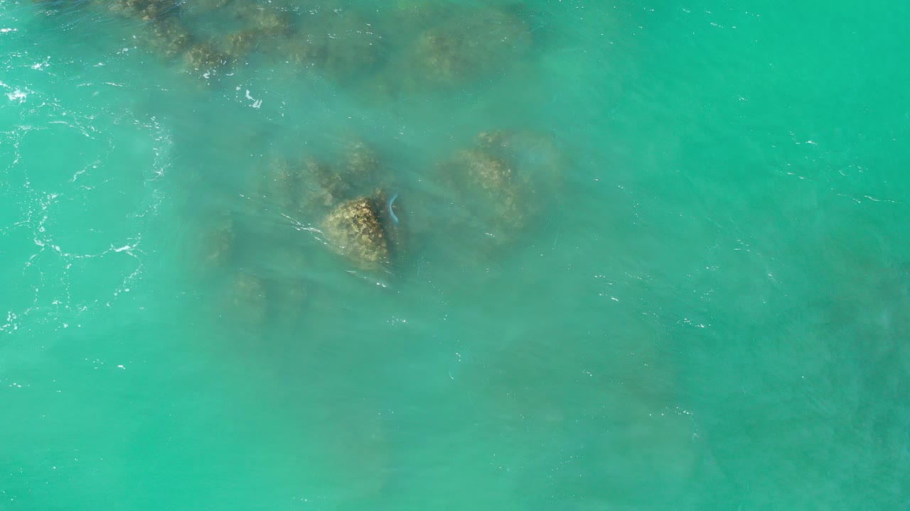 Top-down view of turquoise water in Los Cabos, Mexico, with submerged brown rocks near the upper left and no visible people.