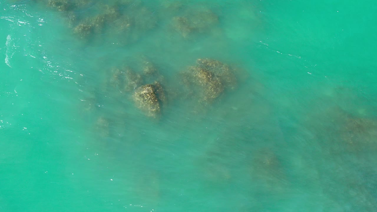 Overhead view of turquoise green water washing over submerged rocks in Los Cabos, Mexico, with small white ripples and no visible people.