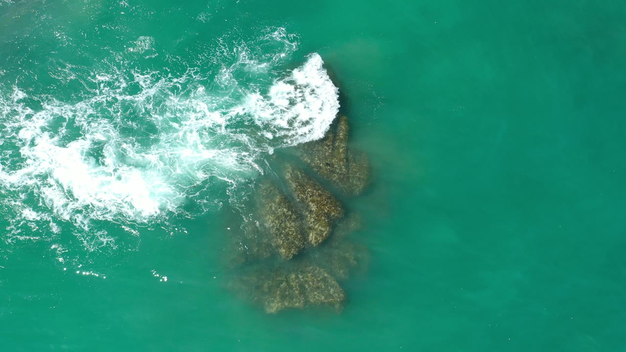 Overhead view in Los Cabos, Mexico, of turquoise sea water and white foam spreading across shallow brown coral rocks, with no visible people.