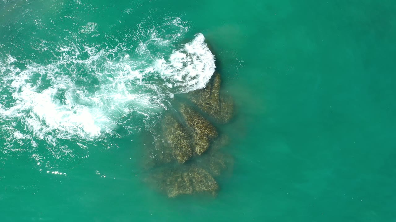Aerial frame from Los Cabos, Mexico, with white surf curling over the upper edge of submerged coral and clear green-blue water around it, with no visible people.