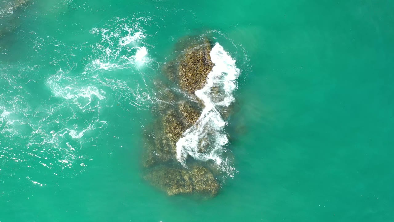 Top-down Los Cabos water detail with a larger rock shape near center right and white foam curling over it, with no visible people.