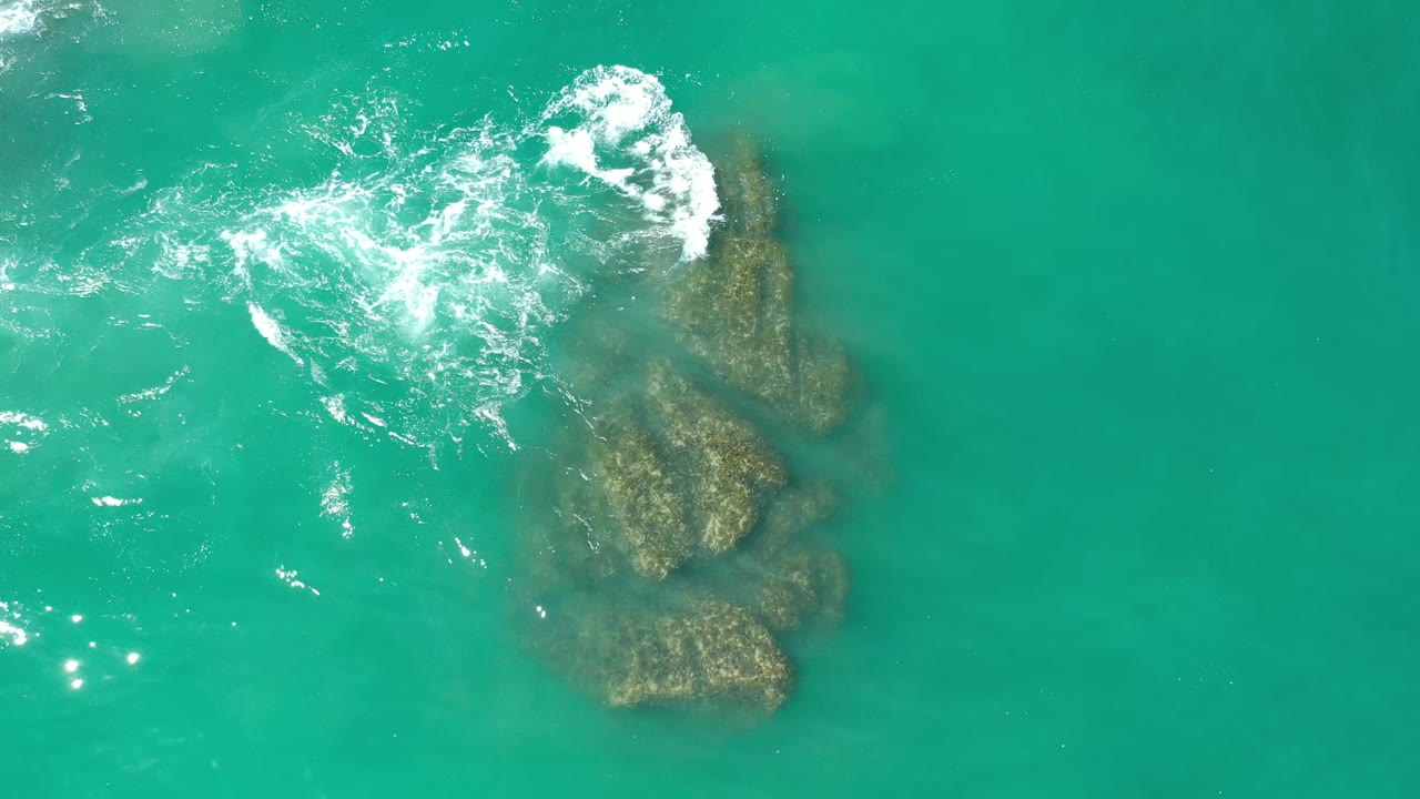 Overhead Los Cabos, Mexico view with the rock cluster slightly right of center, foamy water curling across the upper left side, and no visible people.