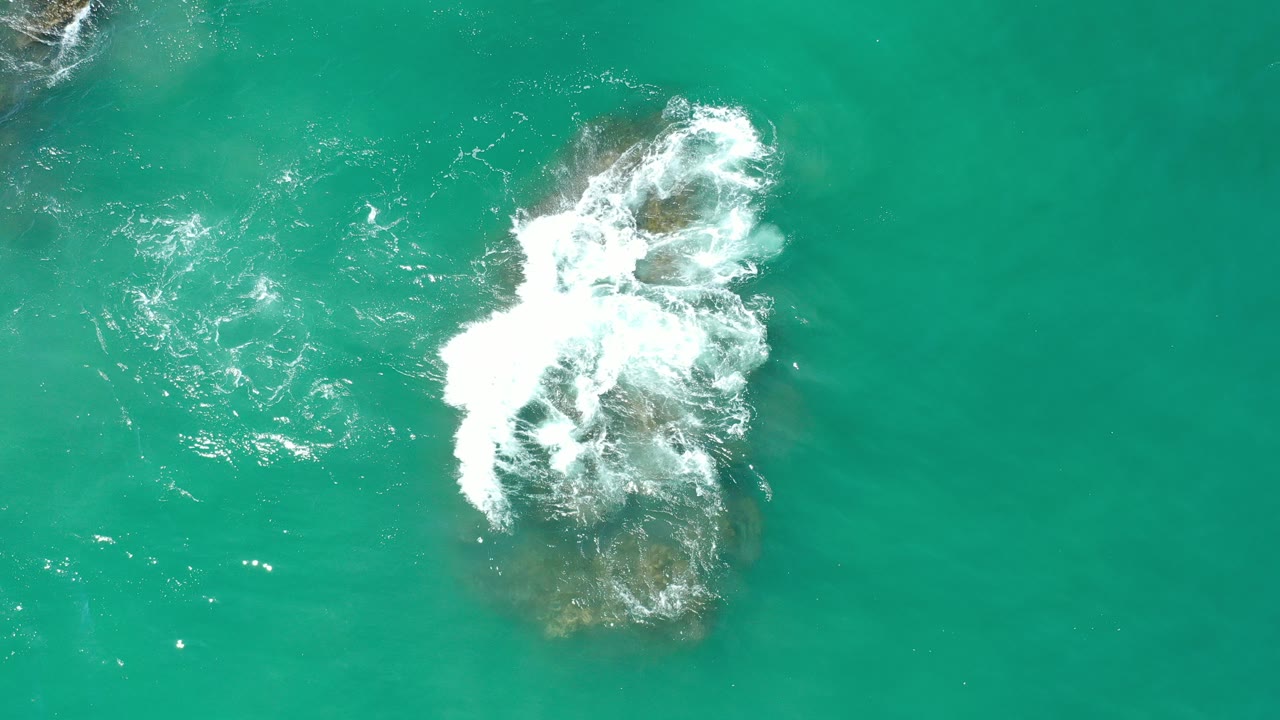 Overhead view in Los Cabos, Mexico of white foam spilling across a brown rock in clear green ocean water, with no visible people.
