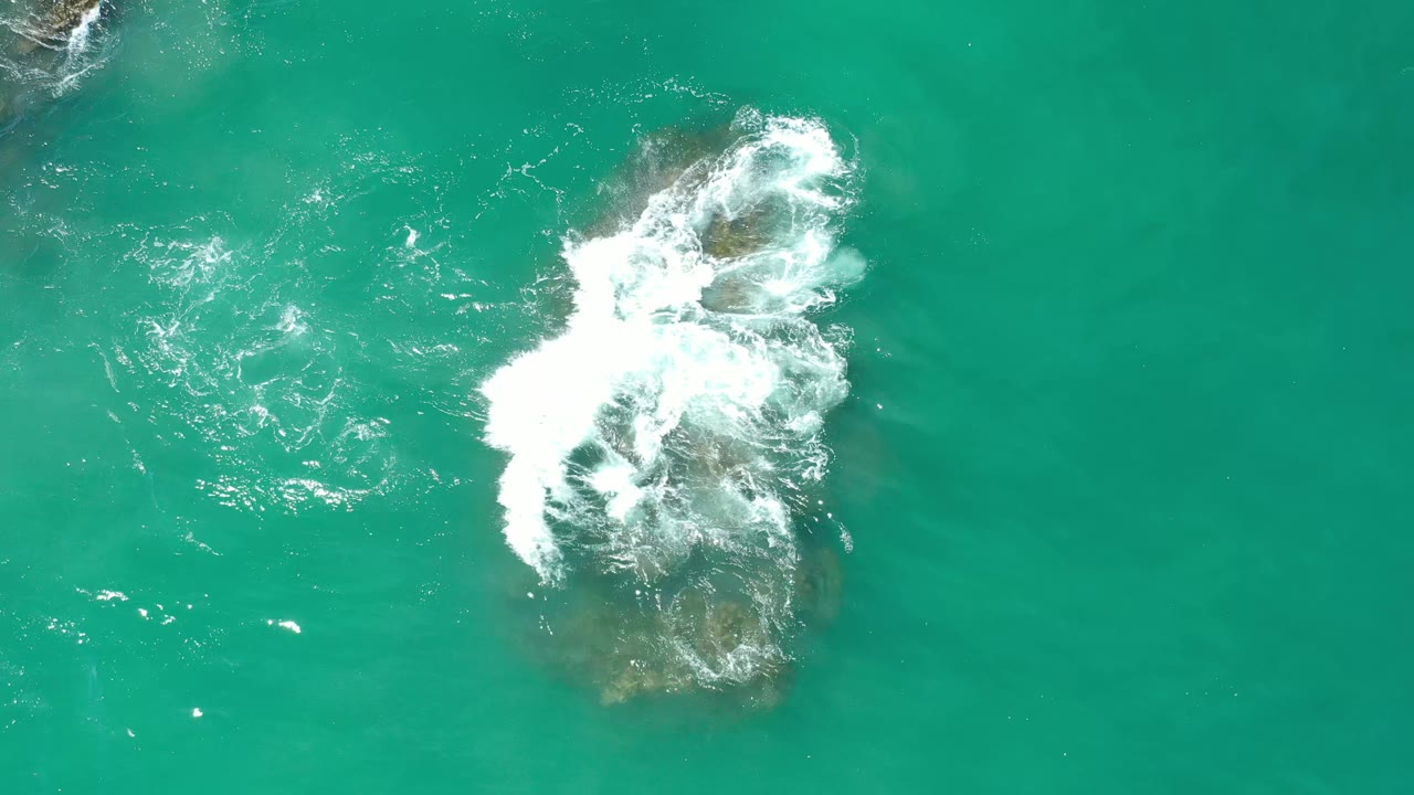 Overhead Los Cabos, Mexico frame with white foam curling over the upper edge of a large brown submerged rock in green water, with no visible people.