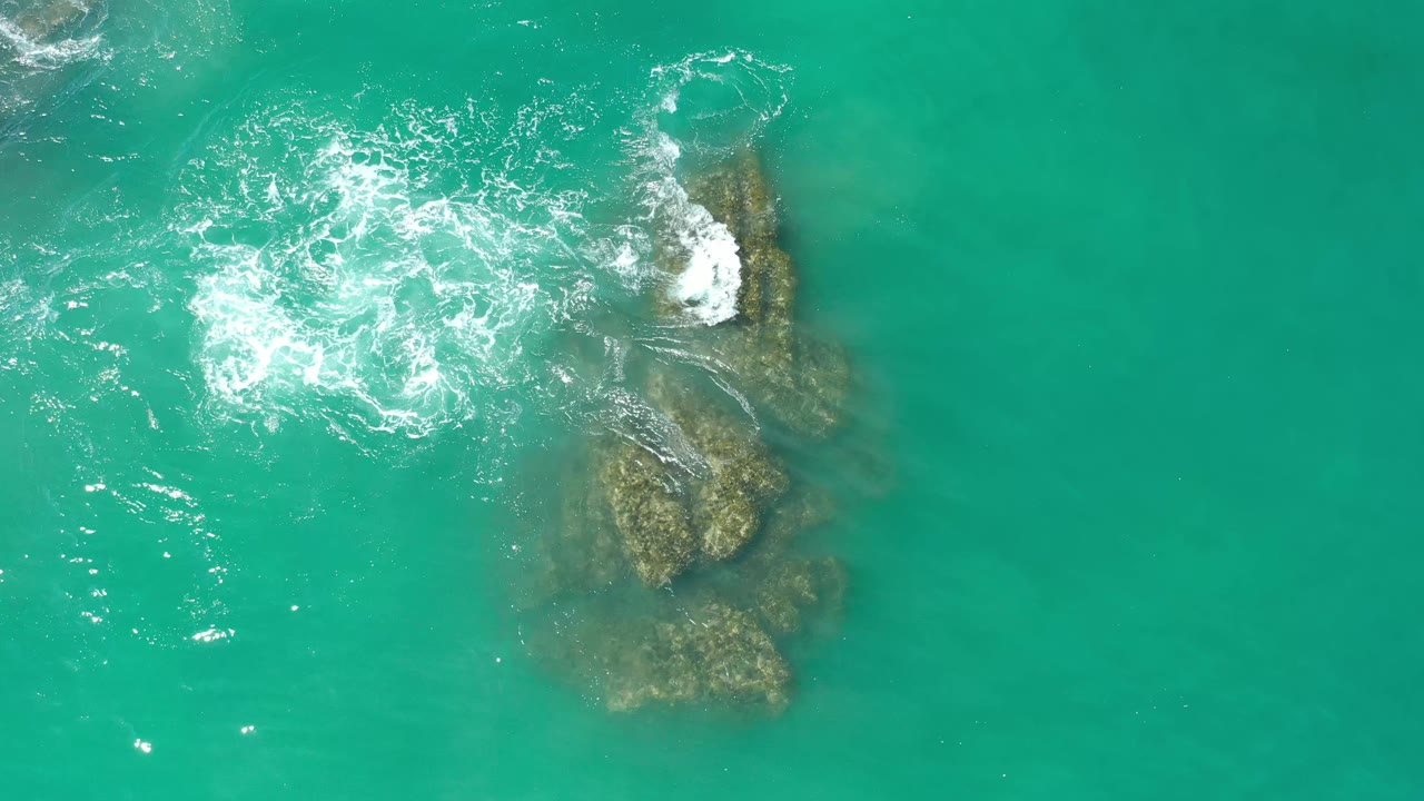 Overhead Los Cabos water detail with foam spreading left of the submerged coral or rock formation and a brighter wave edge along its upper right side, no visible people.