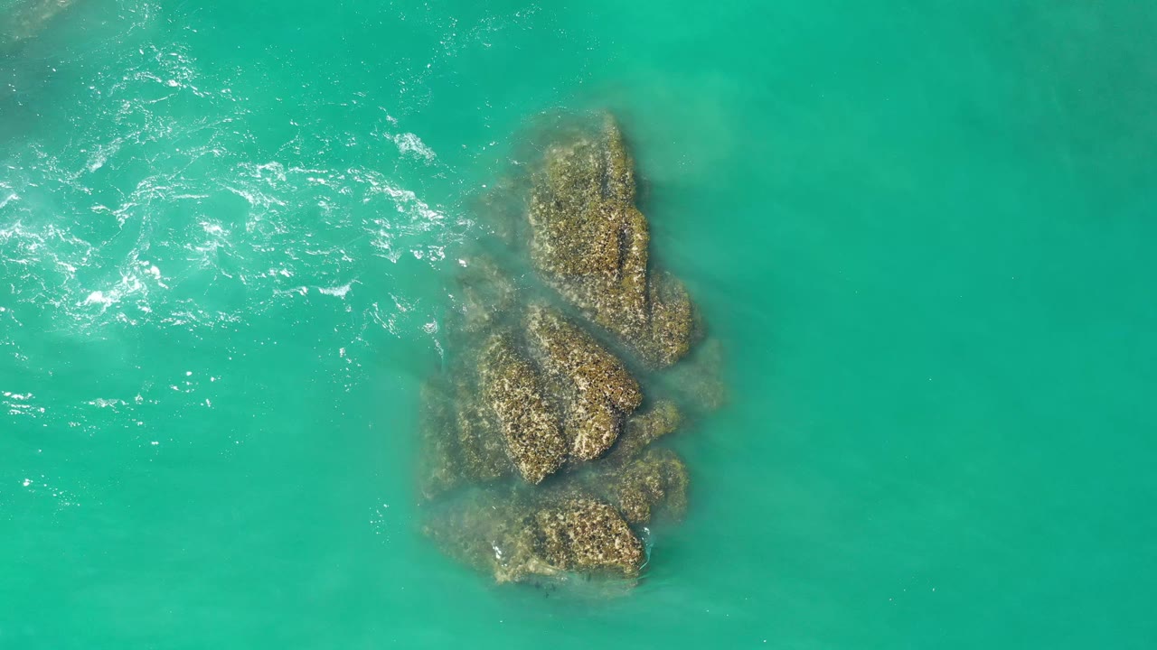 Overhead Los Cabos frame with the rock cluster centered under clear turquoise sea and only light white ripples nearby, with no visible people.
