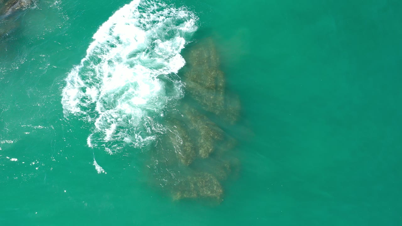 Overhead view in Los Cabos, Mexico of white surf spreading around partly submerged brown beach rocks in turquoise water, with no visible people.