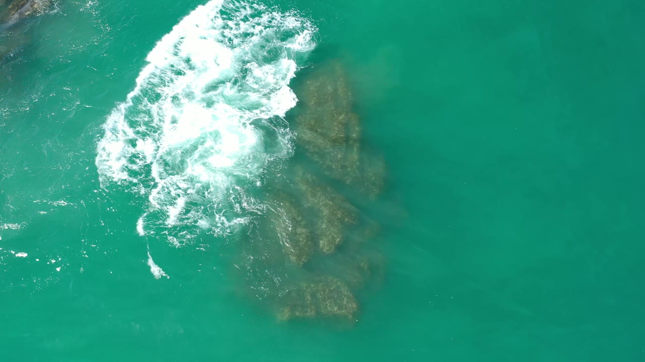 Top-down Los Cabos, Mexico frame with a foamy wave curling beside two submerged rock shapes in green-blue water, with no visible people.