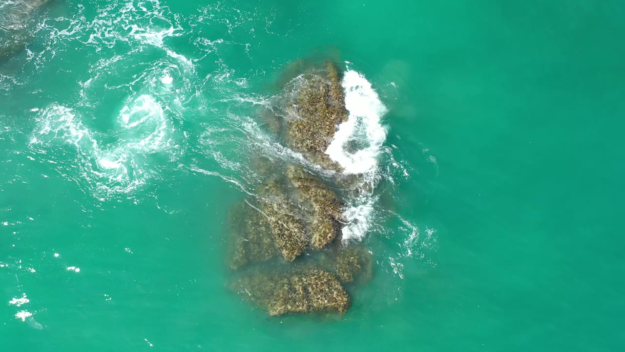 Aerial Los Cabos, Mexico frame with the rock cluster near center right and a separate white foam swirl spreading through clear turquoise water, with no visible people.