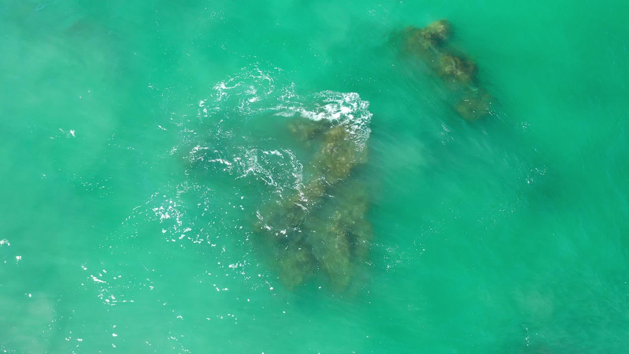 Overhead view of turquoise ocean water in Los Cabos with submerged rocks near the center, bright surface ripples at upper left, and no visible people.