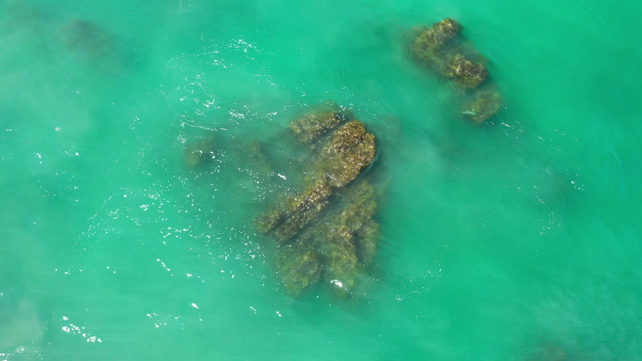 Overhead view of turquoise ocean and dark submerged rocks in Los Cabos, Mexico, with faint ripples and no visible people.