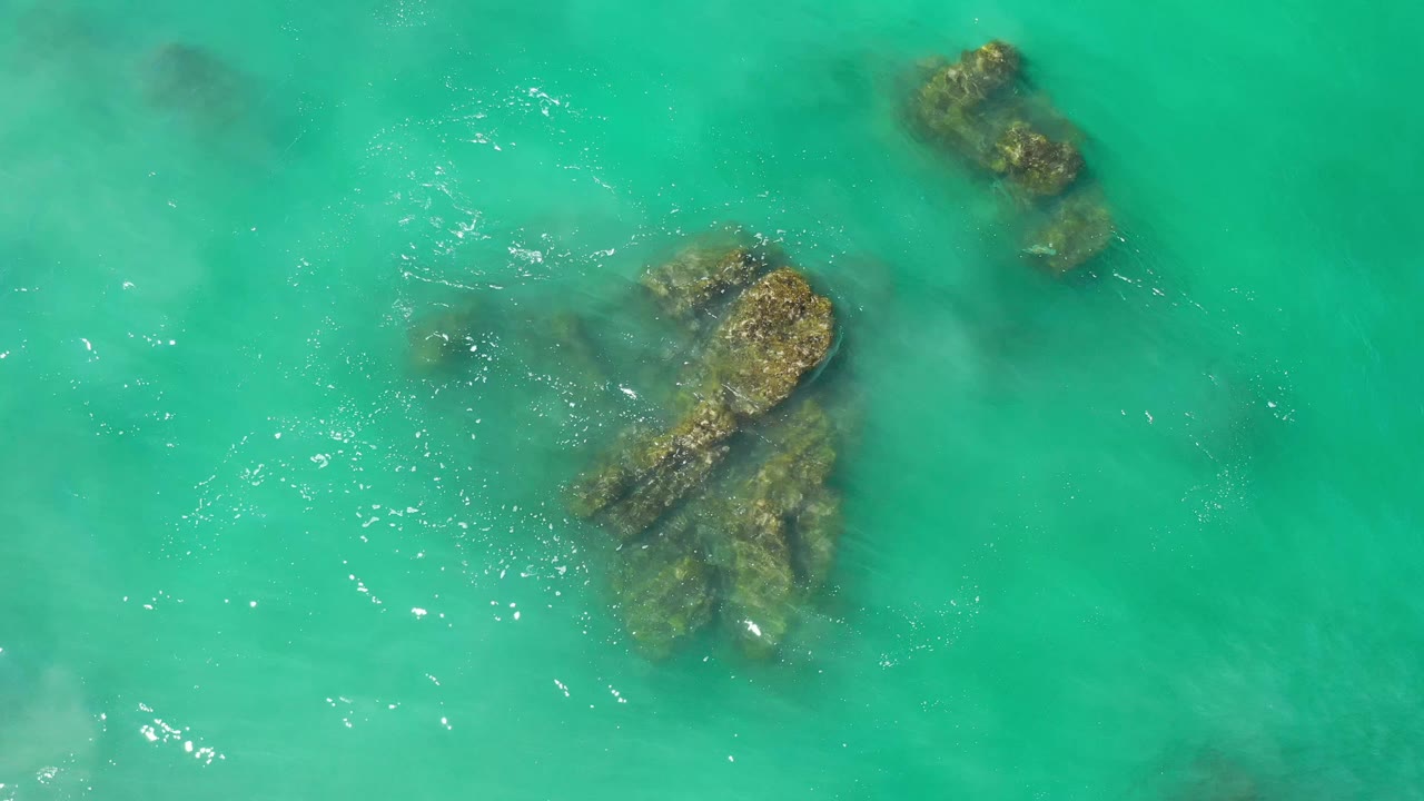 Aerial frame of clear green-blue water over clustered rocks in Los Cabos, Mexico, with small sun glints and no visible people.