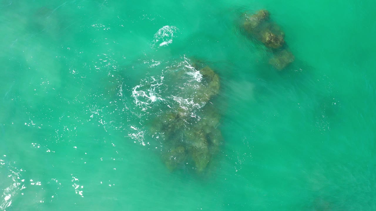 Overhead view in Los Cabos, Mexico, showing turquoise water over two submerged rock formations with small white foam near the lower rock and no visible people.