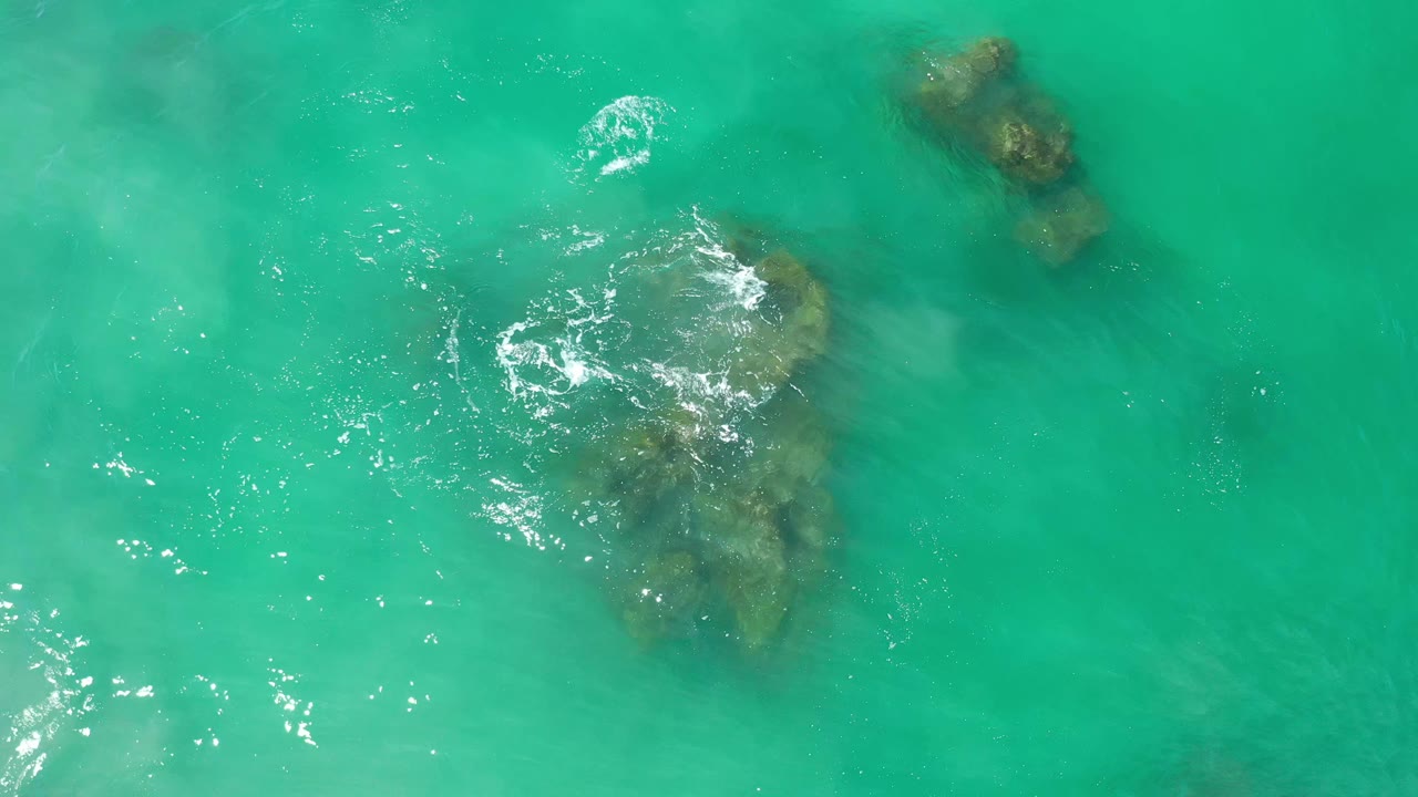 Overhead Los Cabos water view with a larger reef shape near center, a smaller rock near the top, scattered ripples, and no visible people.