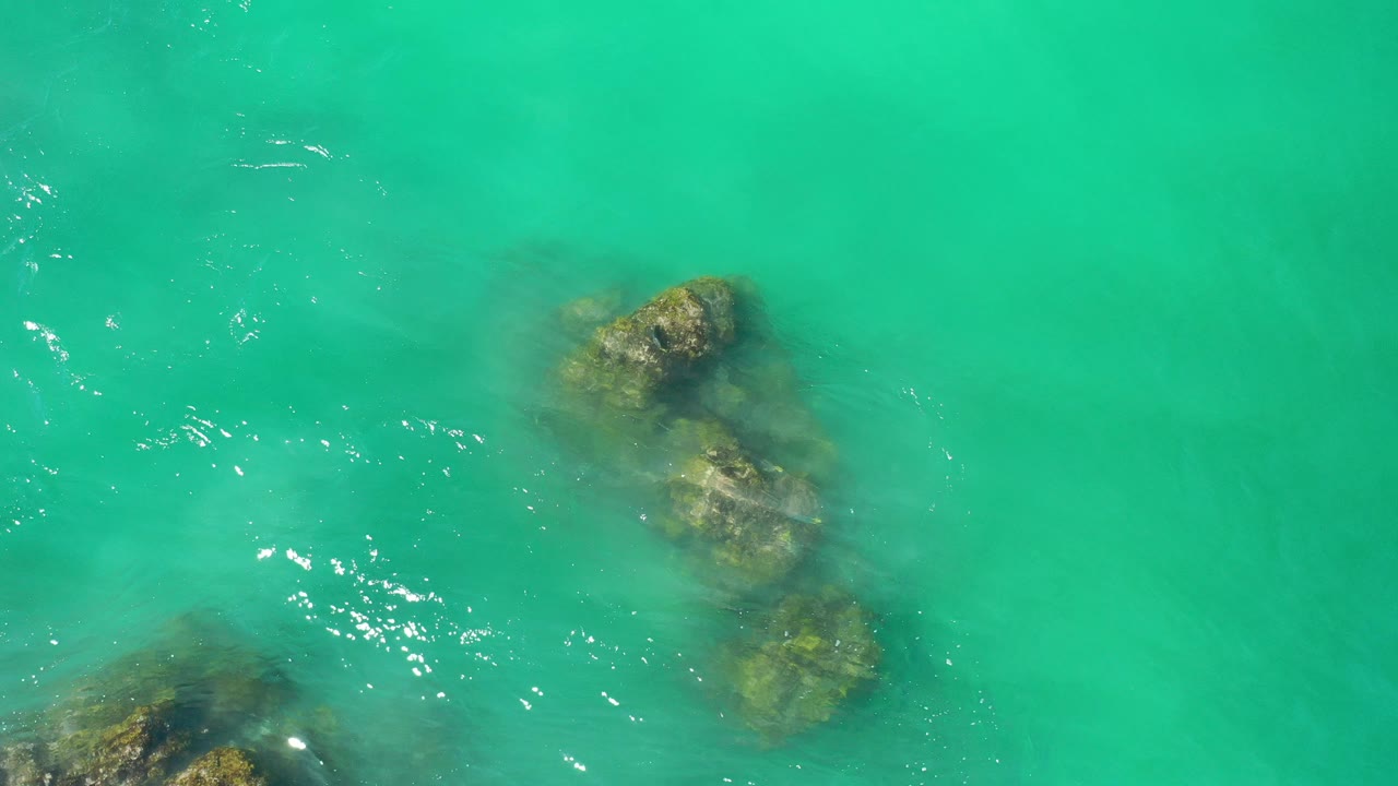 Overhead view in Los Cabos, Mexico, showing green ocean water over several algae-covered rocks near the left and lower edges, with small bright ripples and no visible people.