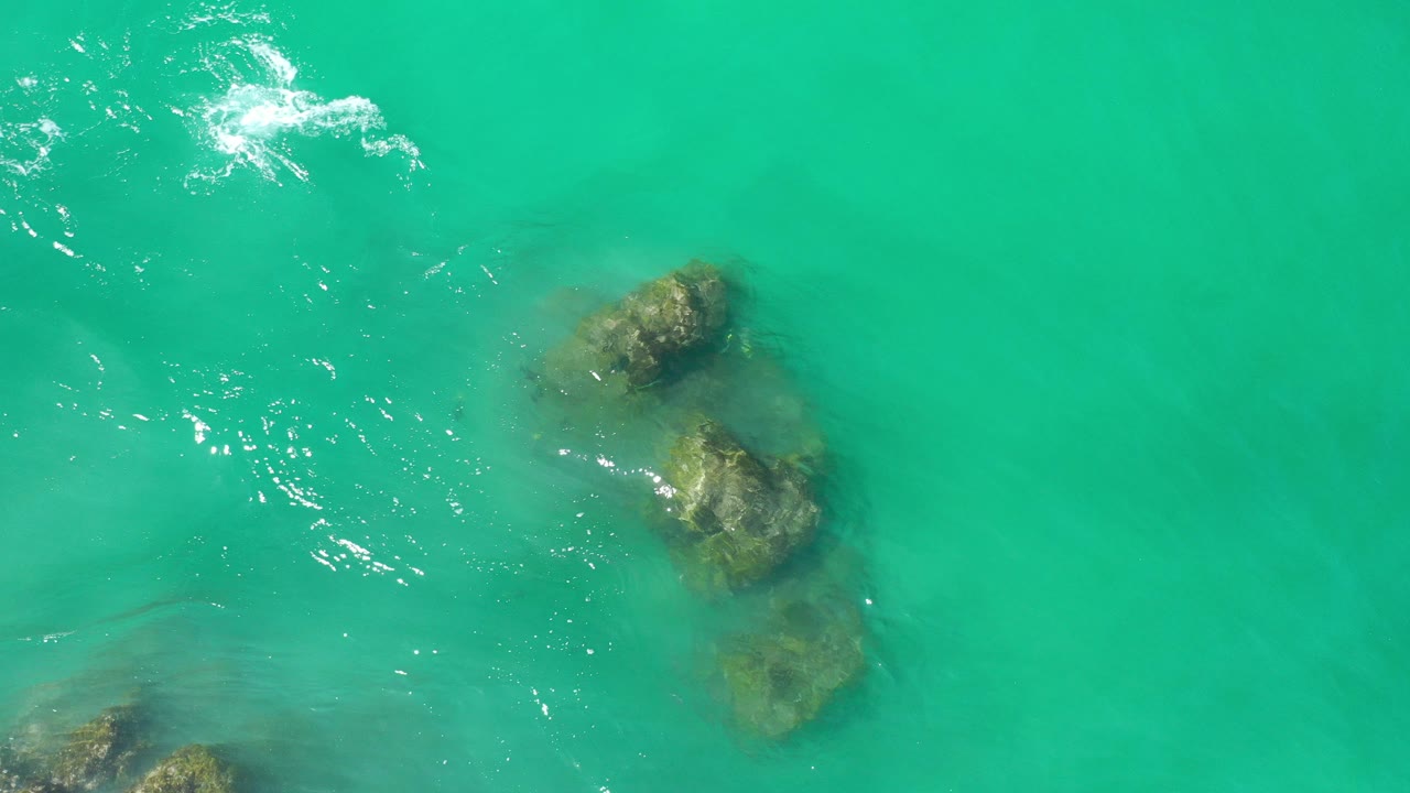Overhead view in Los Cabos, Mexico of turquoise water, a diagonal line of submerged rocks, bright surface glare, and no visible people.